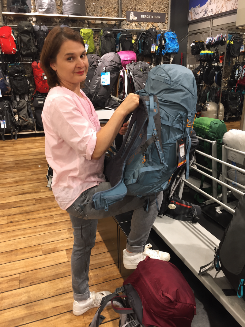 woman in a pink shirt tests a large blue hiking backpack in an outdoor gear store with backpacks on wall racks behind her