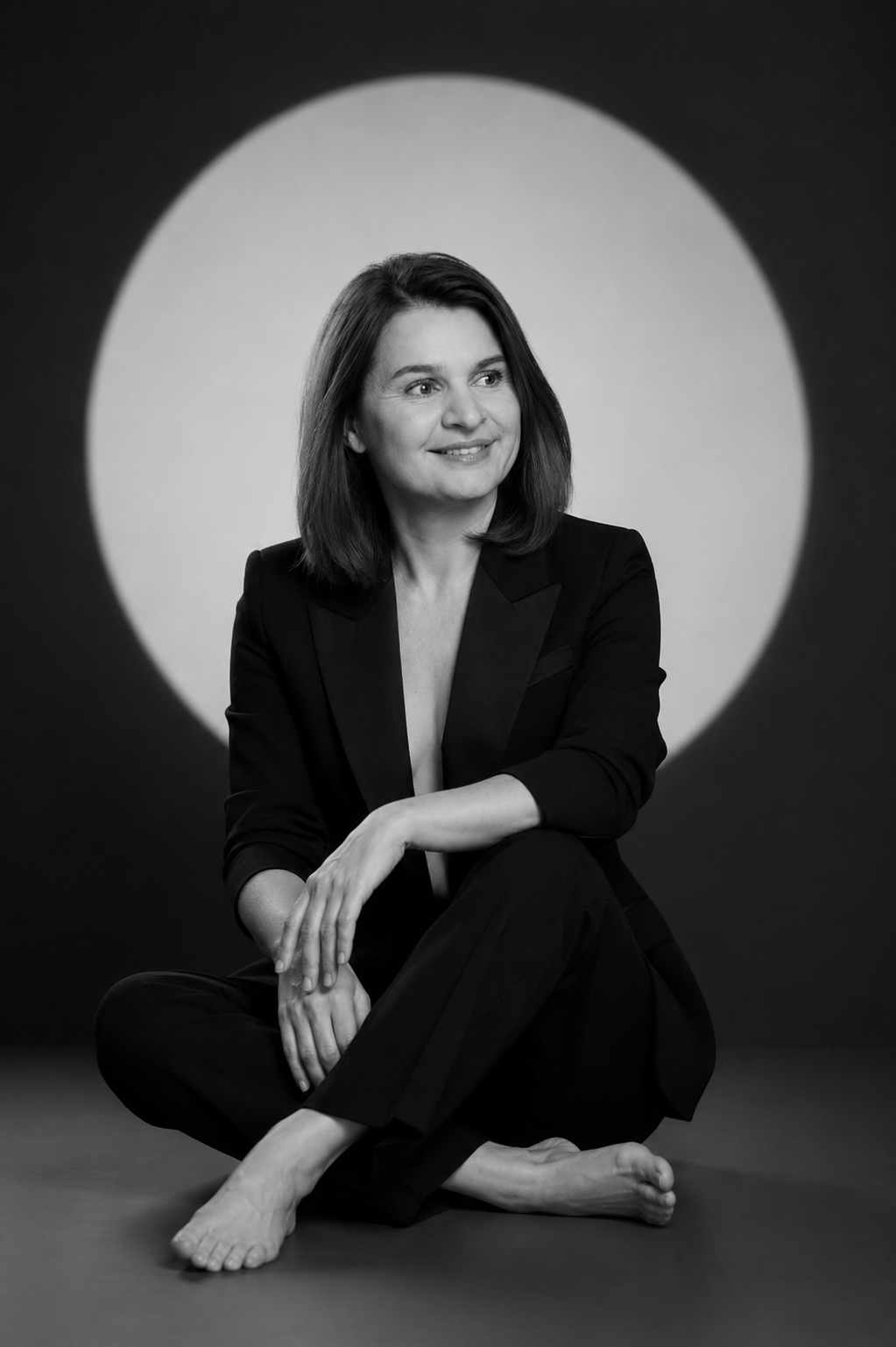 black and white portrait of a woman in a suit sitting cross legged on the floor smiling with a large circular light backdrop behind her