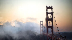 golden gate bridge at sunset shrouded in fog with an orange pink sky above the silhouette of the bridge towers and cables
