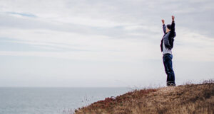 person standing on a grassy cliff by the sea with arms raised in triumph
