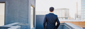 man in a dark business suit stands on a rooftop railing facing a city skyline back view